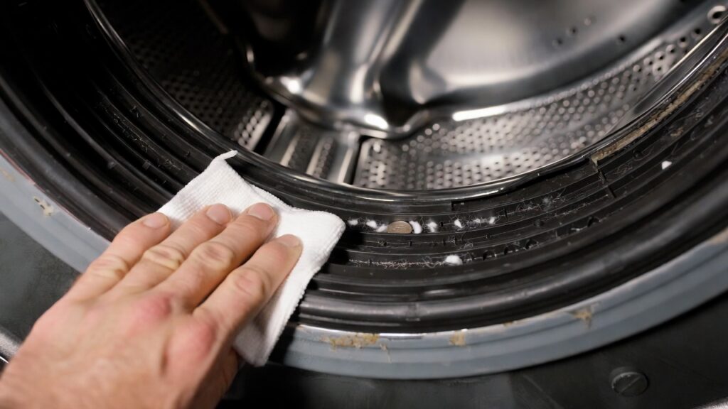 Hand wiping inside the rubber door gasket of a front load washing machine to remove lint and debris for maintenance.