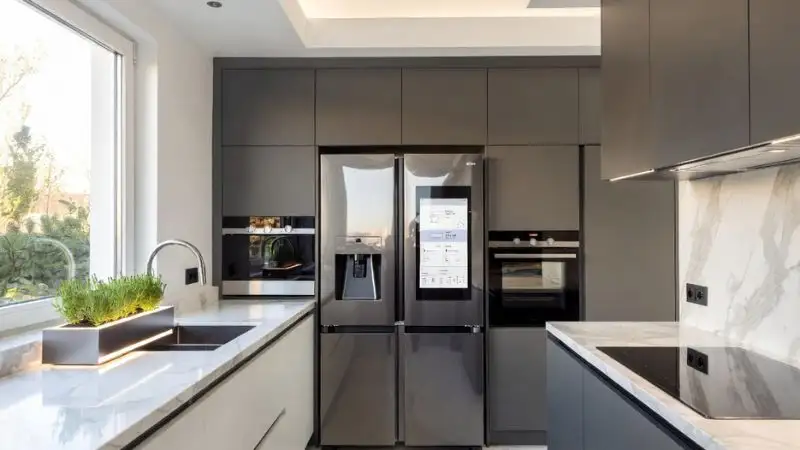 Wide-angle photorealistic shot of a futuristic 2026 smart kitchen filled with natural morning light. Sleek stainless steel AI refrigerator with a transparent glass door displaying a digital shopping list and energy consumption stats. In the background, a smart oven and minimalist induction cooktop integrated into marble countertops. A small elegant countertop herb garden with automated LED lighting near the window. Clean, organized, high-tech yet cozy atmosphere.